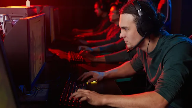 Jogador concentrado, com fones de ouvido, treinando no computador ao lado dos companheiros de equipe em uma sala de jogos iluminada por luzes vermelhas.